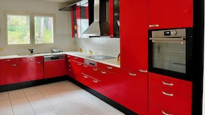 A modern kitchen with red cabinets, a stainless steel oven, and a window with a view of trees. - CAP DE HE' MAIN HOUSE (Lit-et-Mixe)