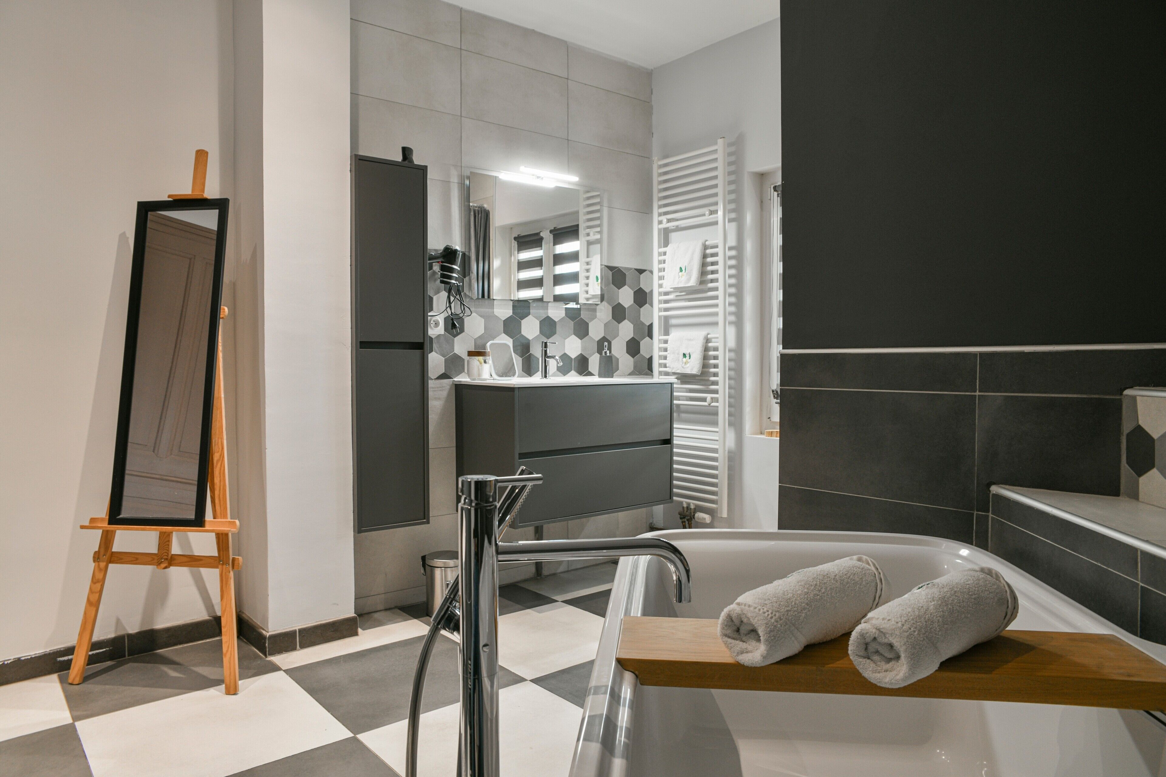 Un baño moderno con piso a cuadros, una bañera, un estante de madera con toallas, un espejo y un perchero para toallas.