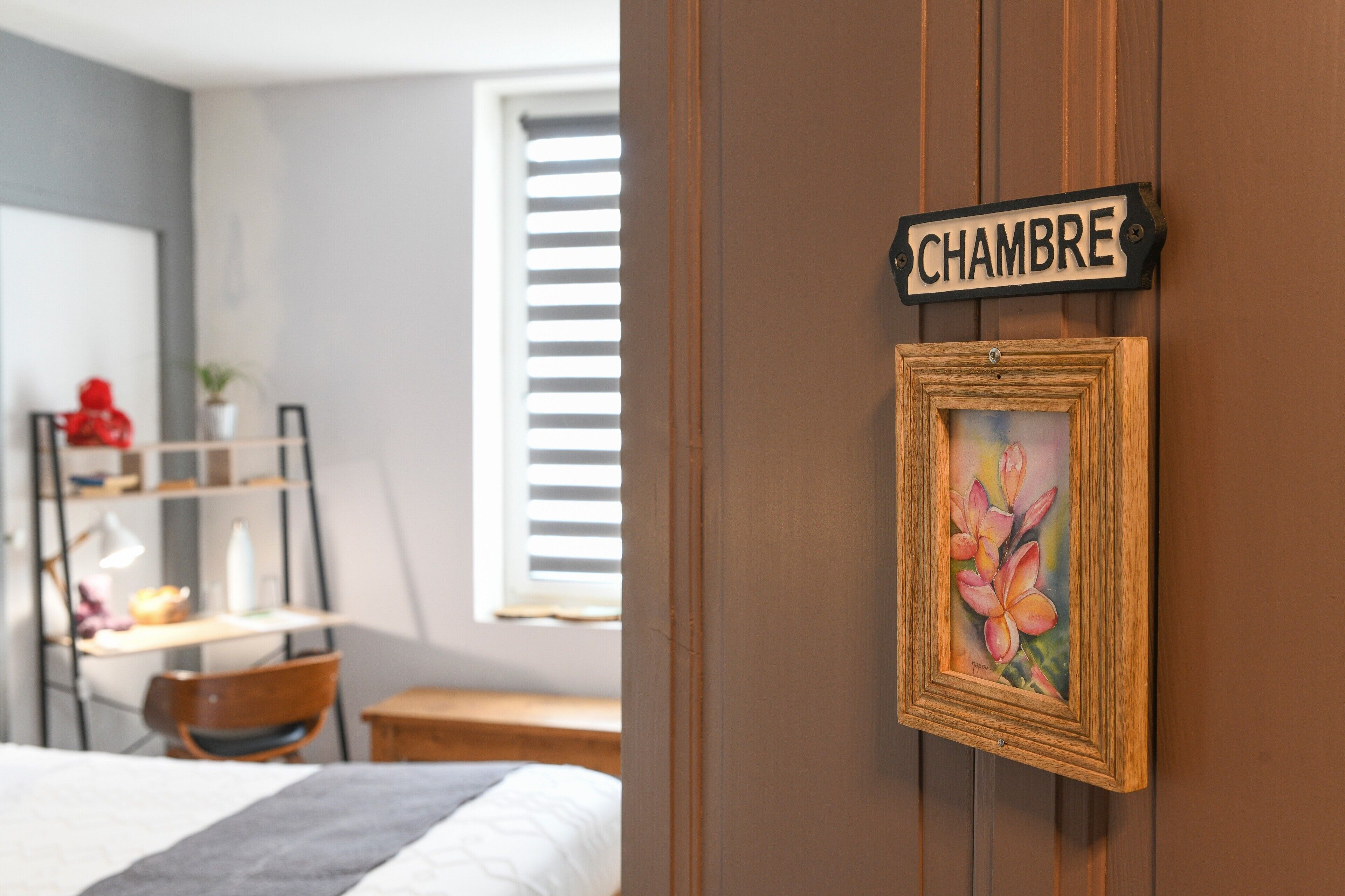 A bedroom with a bed, a desk, a chair, a window with blinds, a door with a sign, and a framed picture.