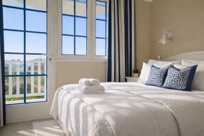 A bedroom with a large bed, white bedding, a view through an open door, and a nightstand with a lamp.
