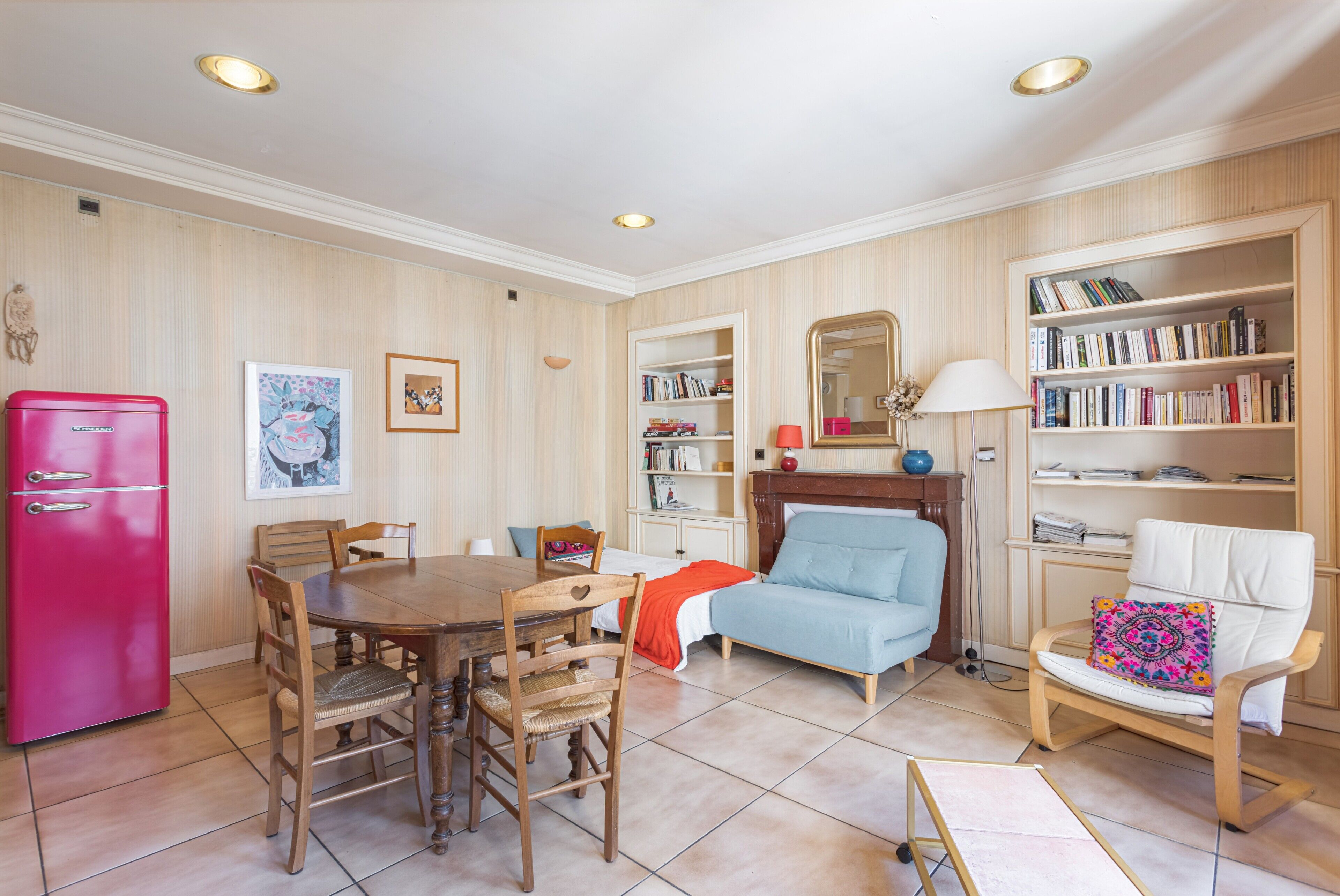 Une pièce comprenant une table à manger, des chaises, une bibliothèque, un canapé, un fauteuil avec un coussin, un miroir et un réfrigérateur rose.