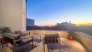 Uma varanda com móveis de exterior, um vaso de planta e vista para o pôr do sol sobre a paisagem urbana.