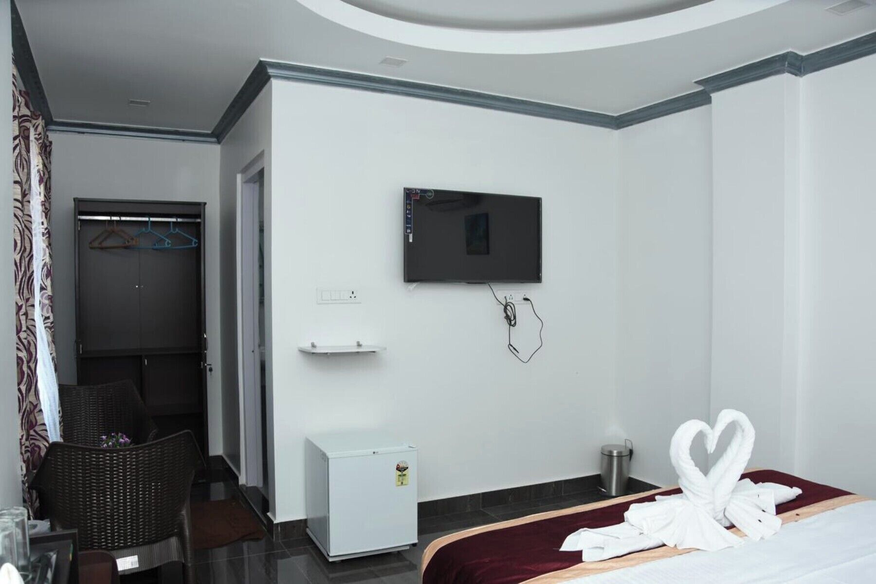 A hotel room with a bed, a TV mounted on the wall, a small white cabinet, and a wicker chair.