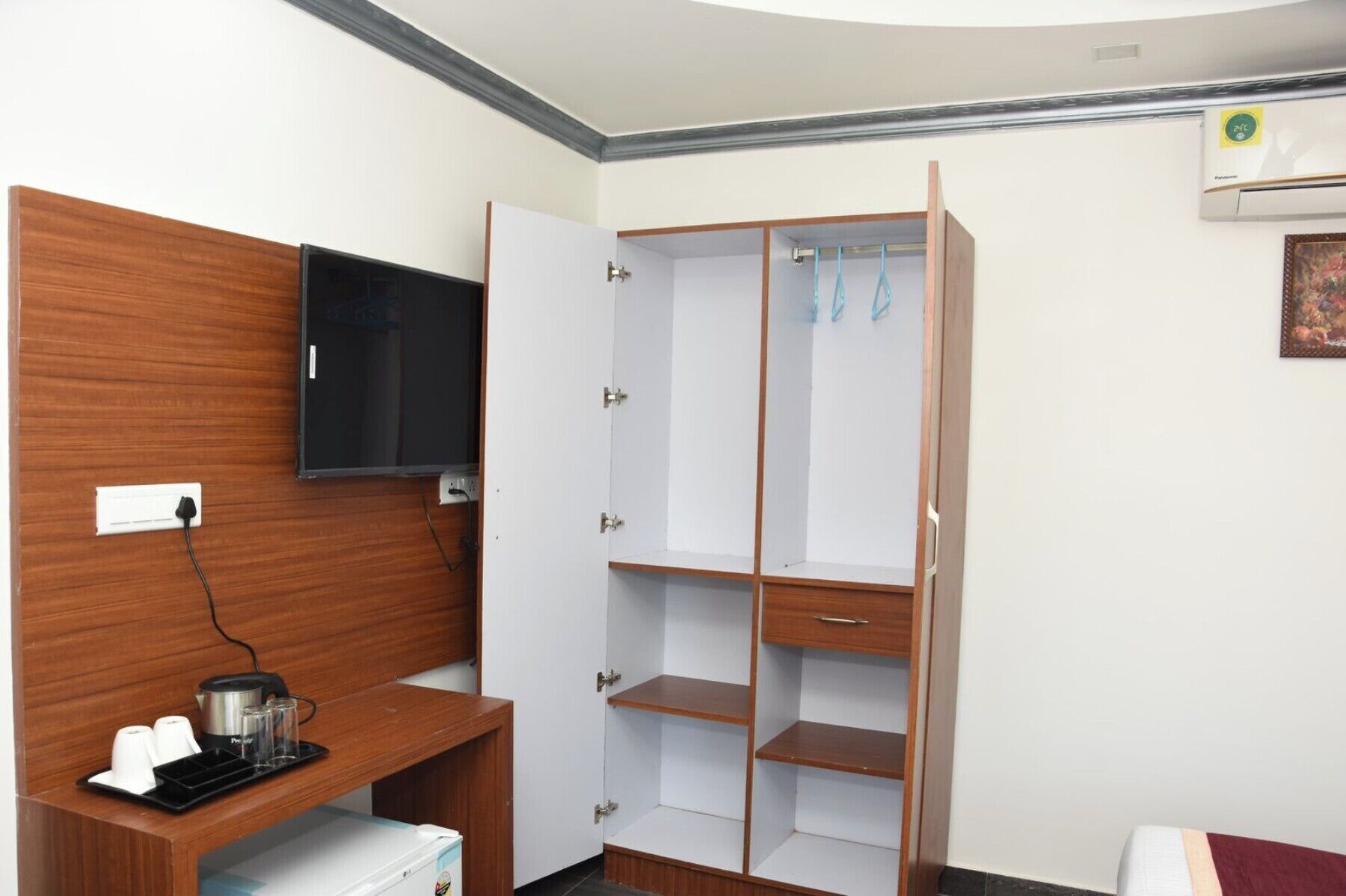 A hotel room with a wooden desk, a television, a wardrobe, and a small refrigerator.
