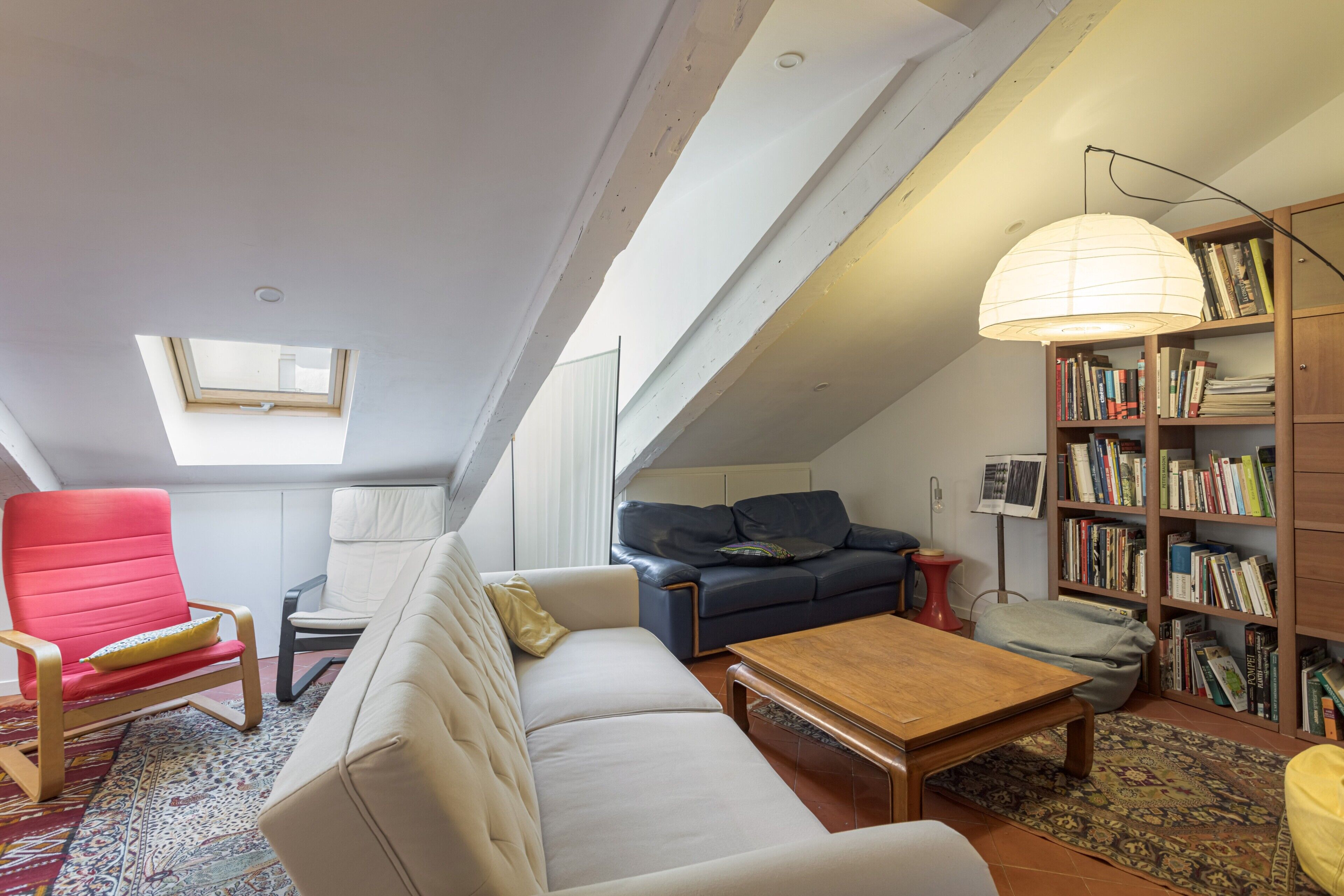 Une pièce de style « loft » avec un plafond en pente, une lucarne, un canapé, un fauteuil rouge, une table basse en bois, une bibliothèque et une lampe.