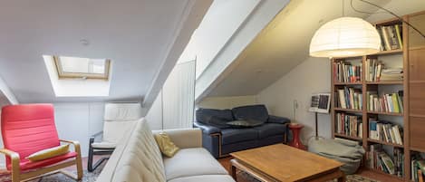 Une pièce de style « loft » avec un plafond en pente, une lucarne, un canapé, un fauteuil rouge, une table basse en bois, une bibliothèque et une lampe.