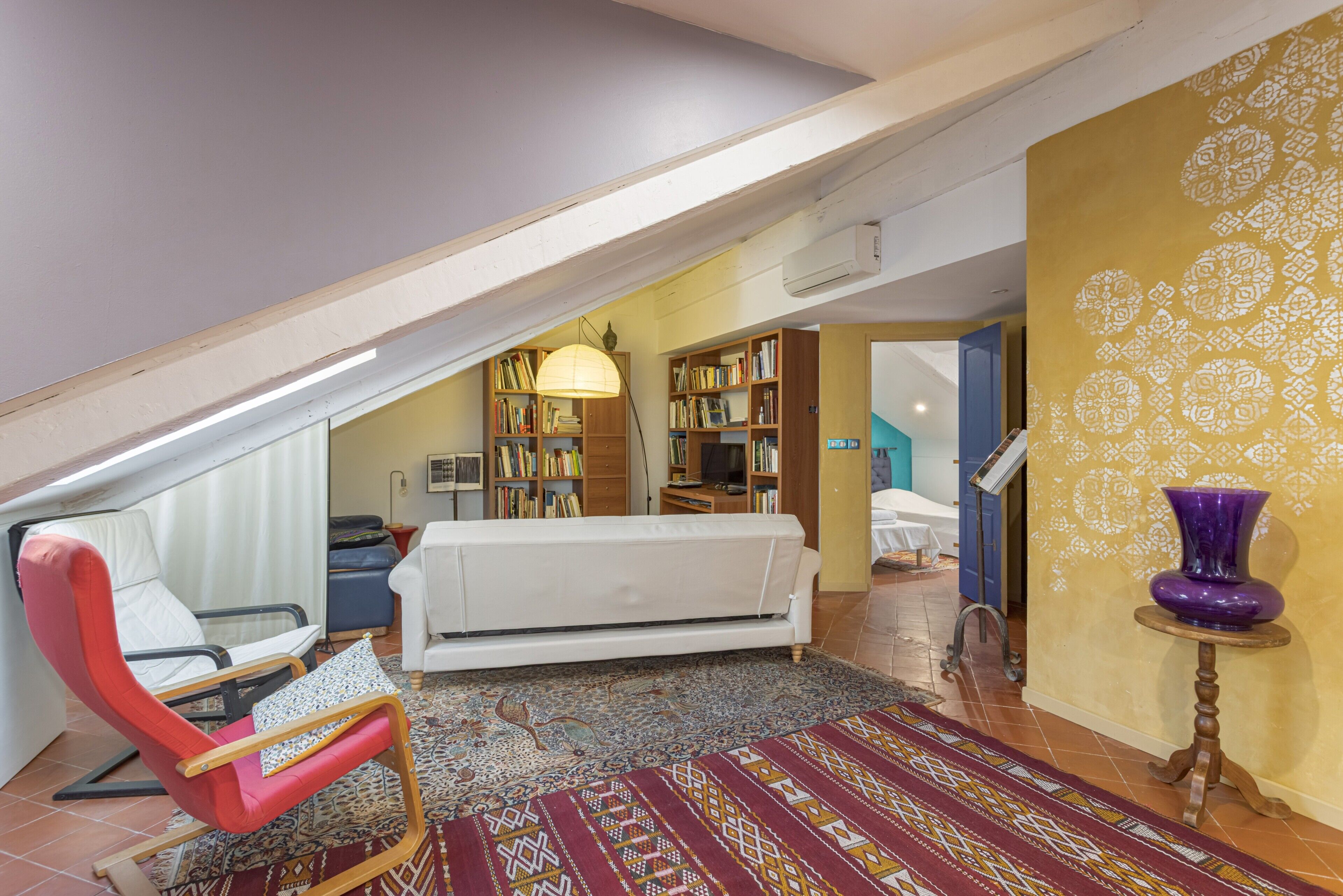 A loft-style room with a high ceiling, a white sofa, a red chair, a patterned rug, and a bookshelf.