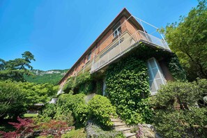 A two-story house with a balcony, surrounded by lush greenery and mountains in the background.