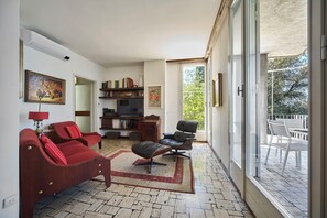 A living room with a sofa, armchair, and a TV. There is a bookshelf, a rug, and a balcony with outdoor furniture.