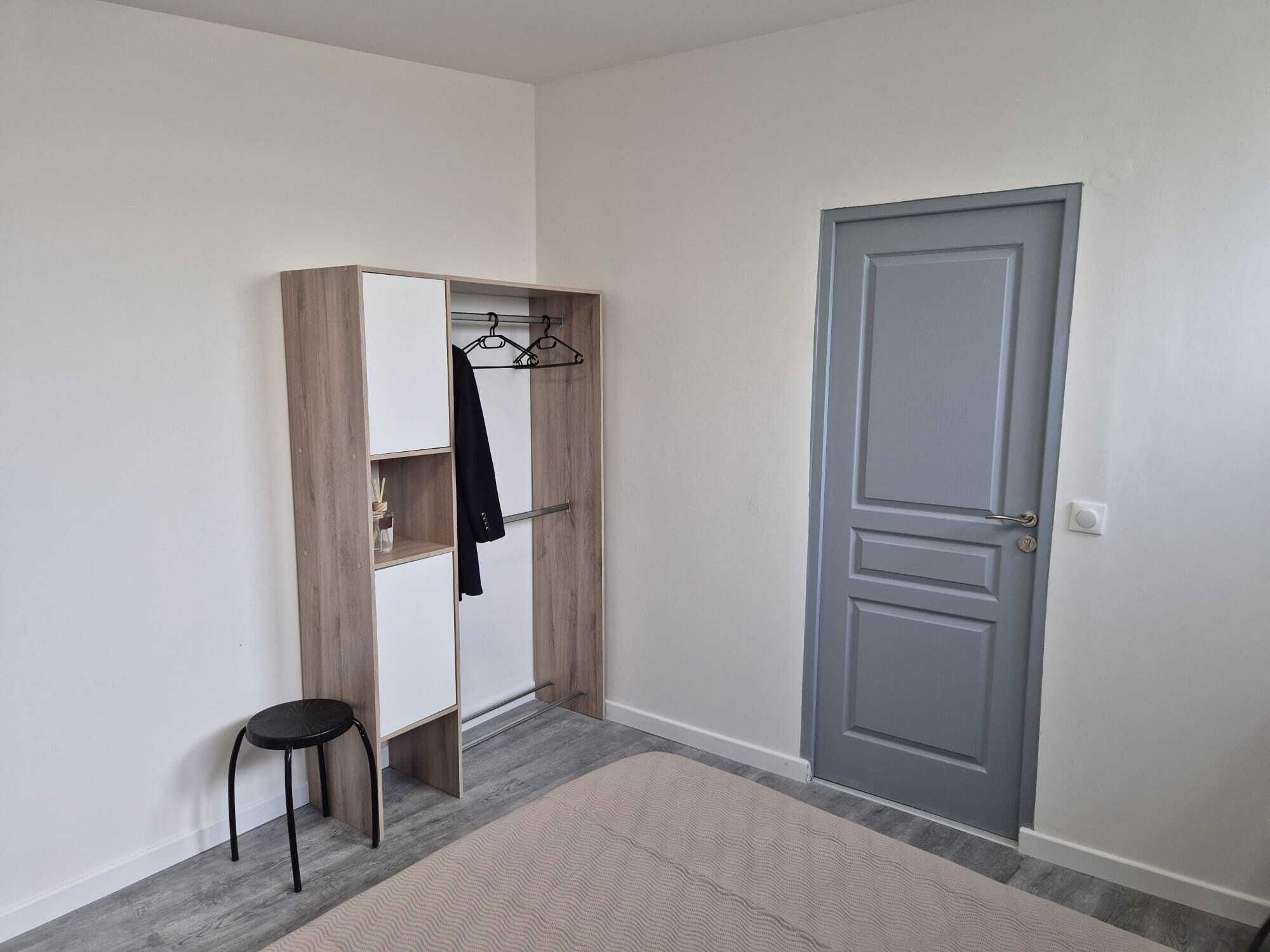 A room with a grey door, a wooden wardrobe with a hanging rack, and a small stool.