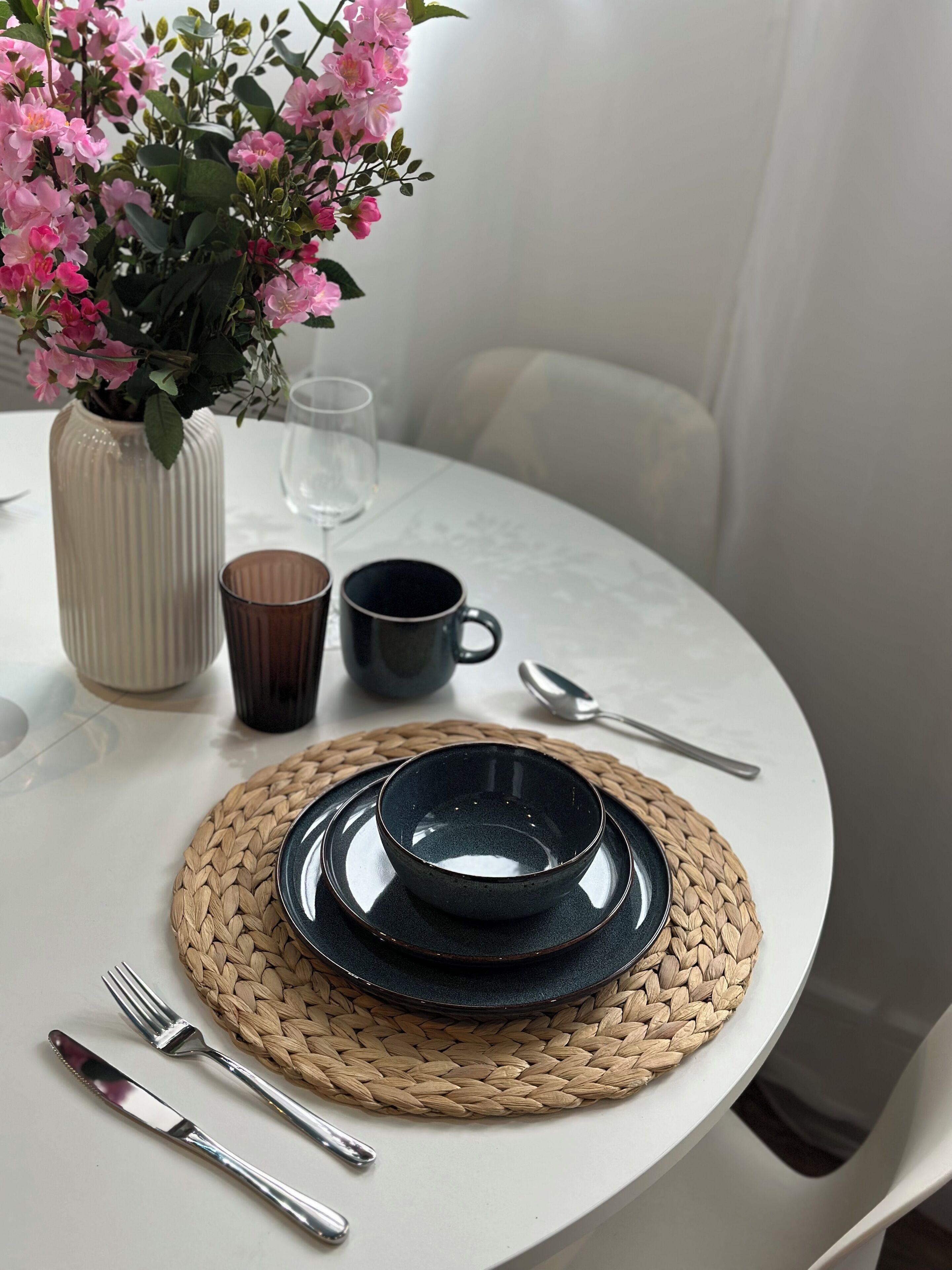 A round table with a woven placemat, a dark bowl, a cup, a glass, and cutlery. A vase with pink flowers is on the table.