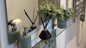 A bathroom with a glass shelf holding a potted plant, two bottles of liquid soap, and a black diffuser. A towel rack and a mirror are also visible. - Beautiful studio flat just 3 minutes walk from Tottenham Spur Stadion! (London)