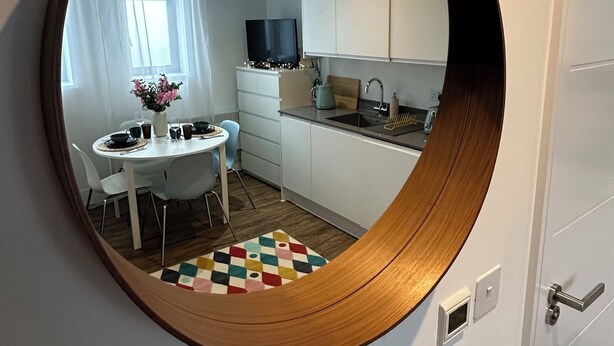 A round mirror on a wall reflecting a kitchen and dining area with a white table, chairs, and a colorful rug. - Beautiful studio flat just 3 minutes walk from Tottenham Spur Stadion! (London)