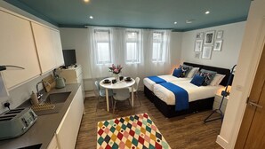 A compact hotel room with a kitchenette, a dining table with chairs, a bed with a blue and white duvet, and a colorful rug. - Beautiful studio flat just 3 minutes walk from Tottenham Spur Stadion! (London)