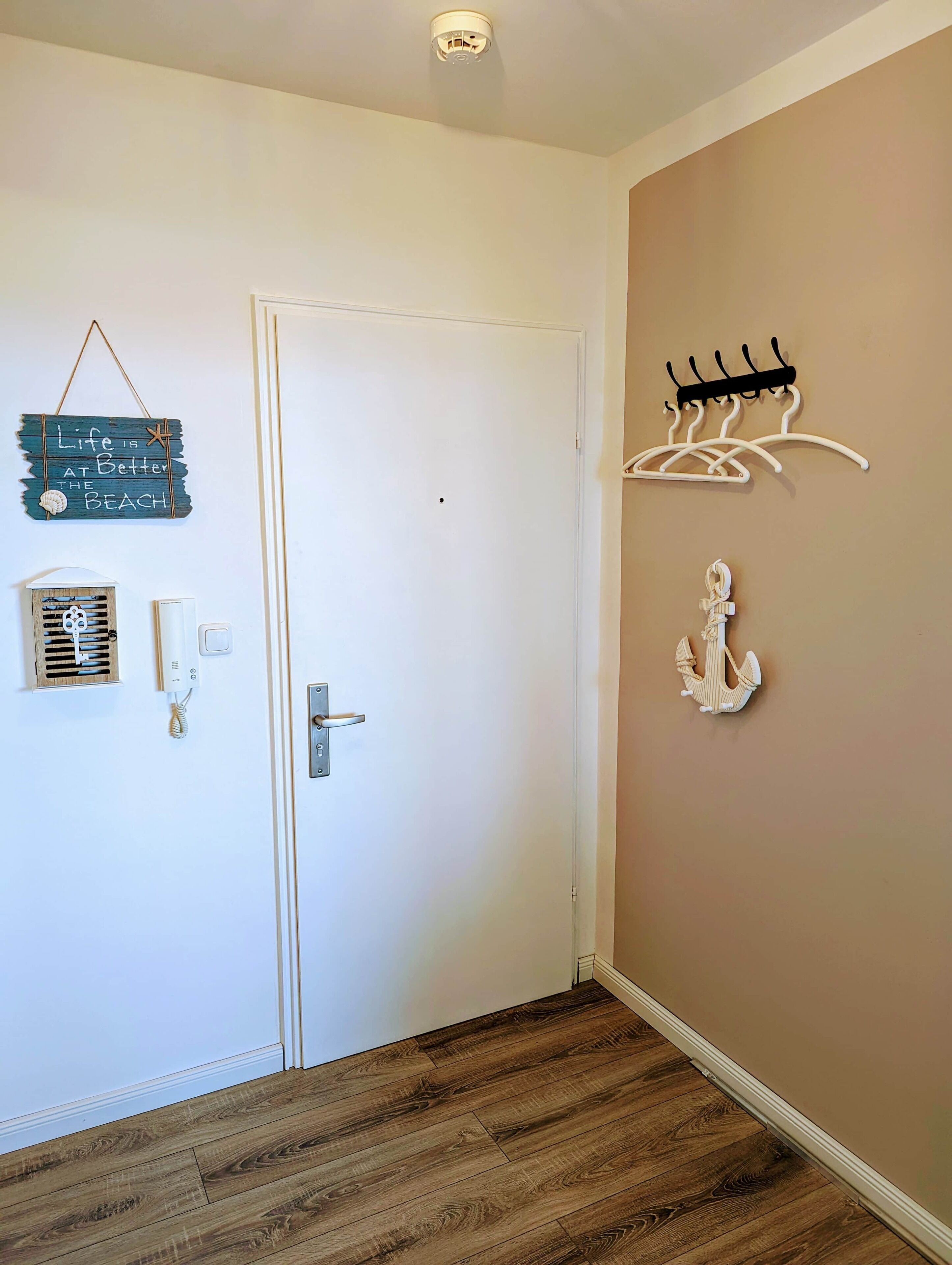 A hallway with a white door, a wall-mounted heater, a wall-mounted phone, a small wooden vent, a wall-mounted coat rack, and a wall-mounted anchor.