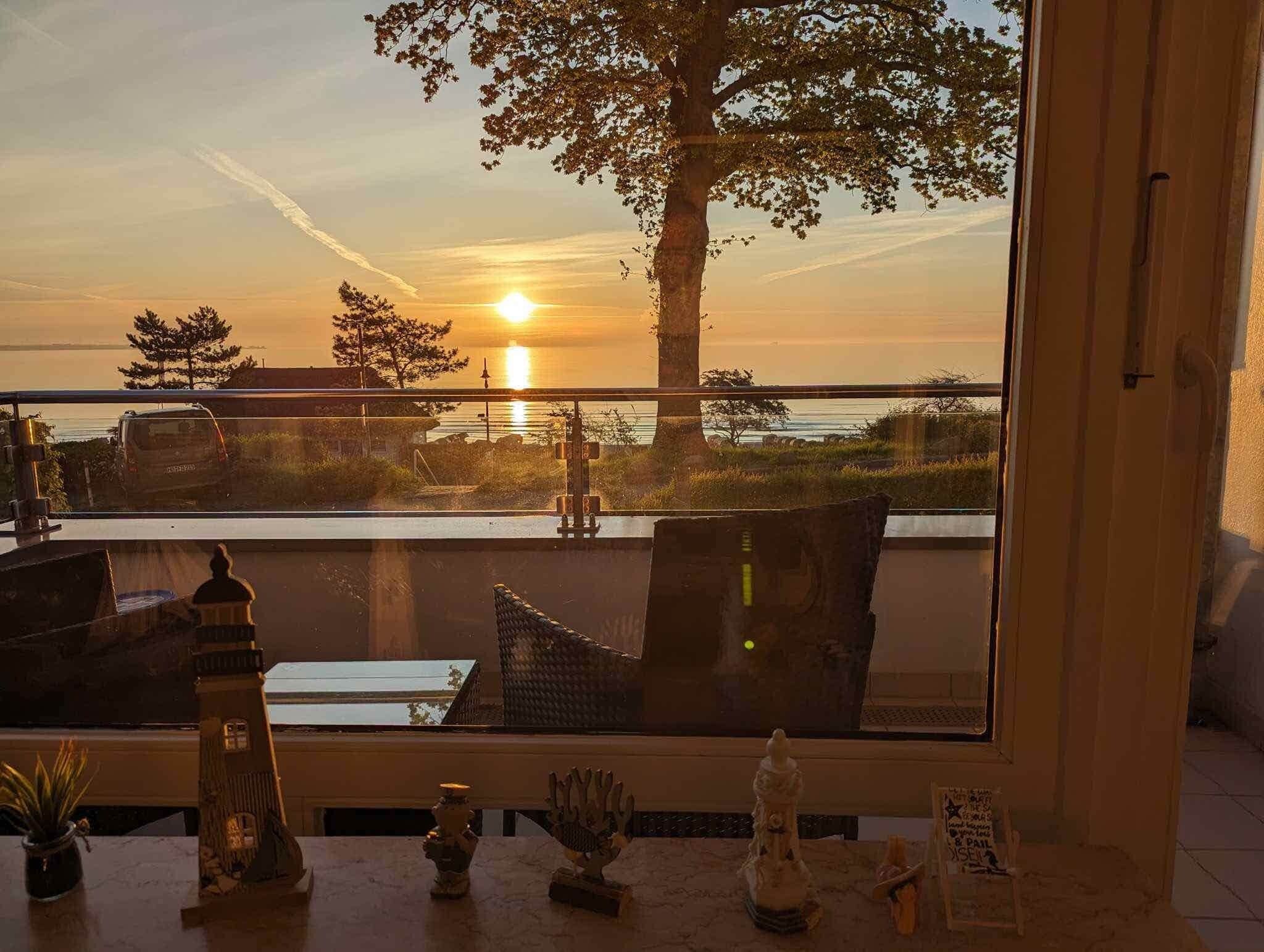 Vista attraverso una finestra di un tramonto su un corpo d'acqua, con un albero e un edificio in lontananza.