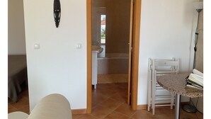 A room with a door leading to a bathroom, a small table with a lamp, and a bed in the background. - Cantinho da Luísa (Coimbrã- Atouguia da Baleia)
