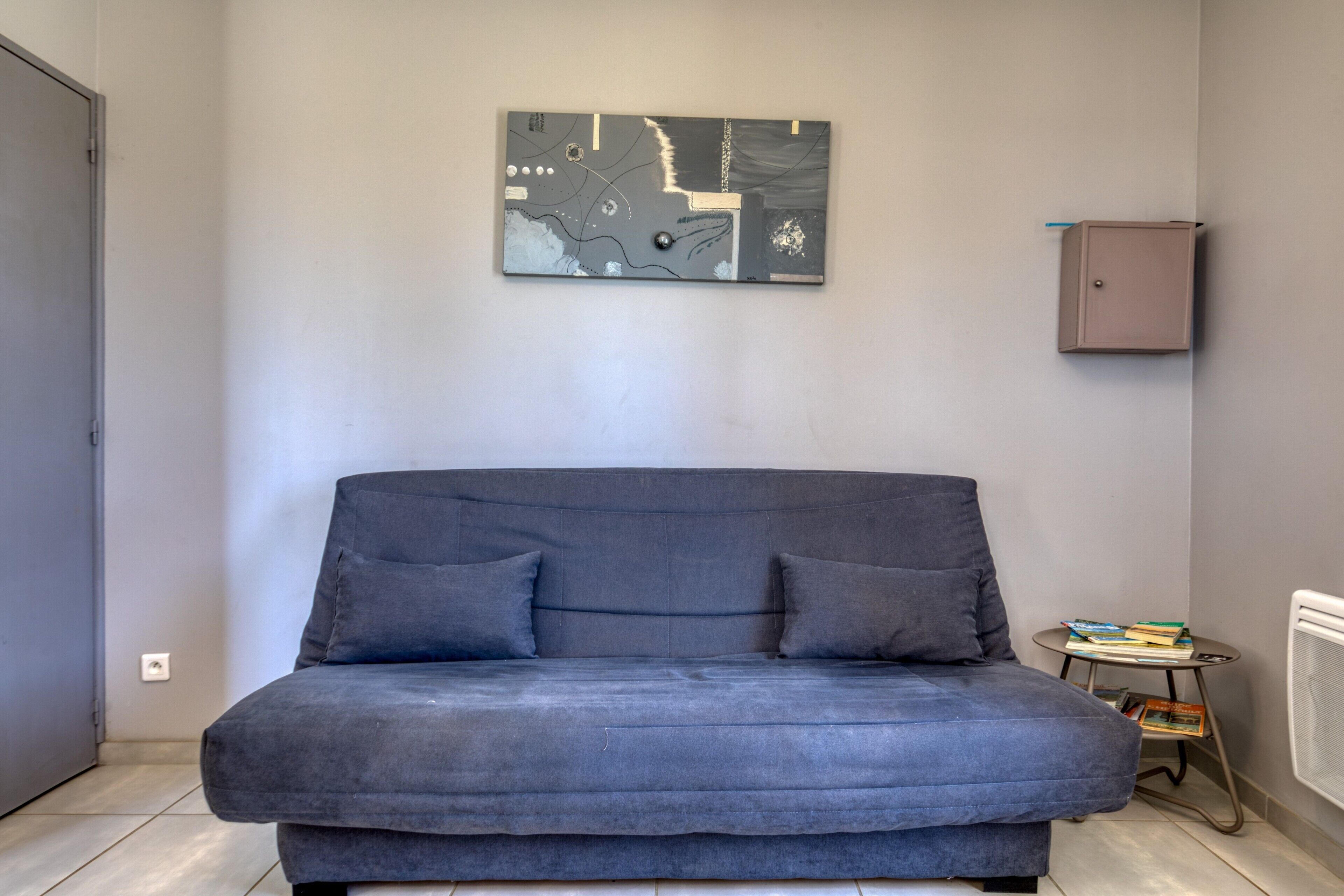 Een grijze bank met twee kussens, een aan de muur hangend abstract kunstwerk, een klein metalen tafeltje met boeken en een radiator.