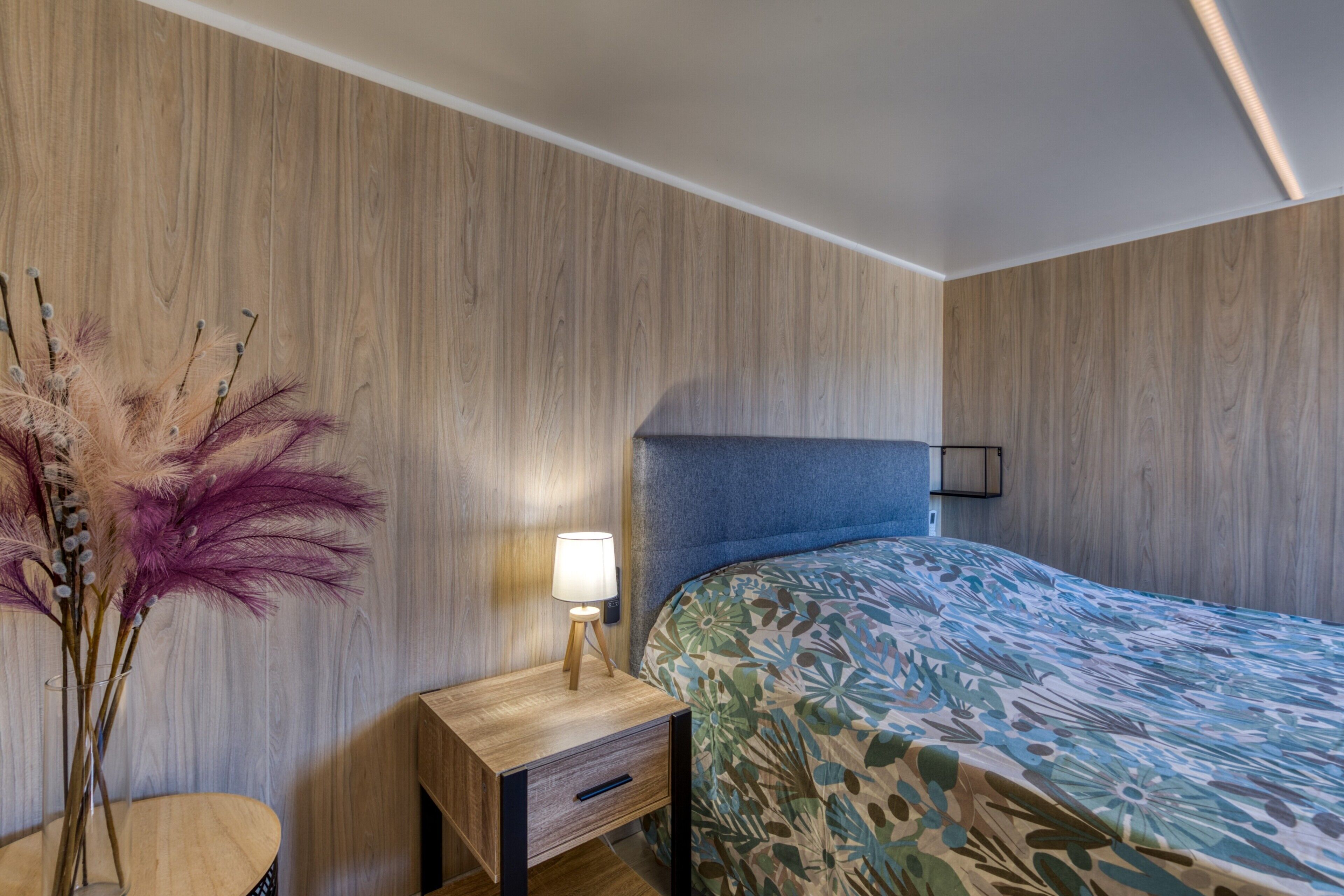 A bedroom with a wooden wall, a bed with a floral bedspread, a bedside table with a lamp, and a vase with dried flowers.