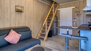 Un espace de vie compact avec un mur en bois, un canapé agrémenté d’un coussin rose, une échelle en bois, un coin cuisine avec un réfrigérateur et un tabouret de bar.