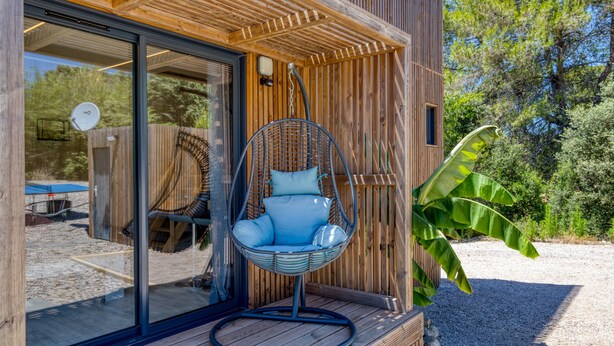 Une façade en bois avec une porte en verre, un fauteuil suspendu et une grande plante.