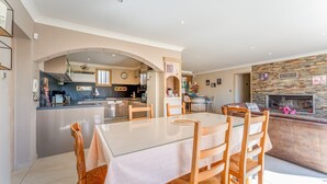 A modern kitchen with a central island, dining area with a glass table and wooden chairs, and a stone fireplace.