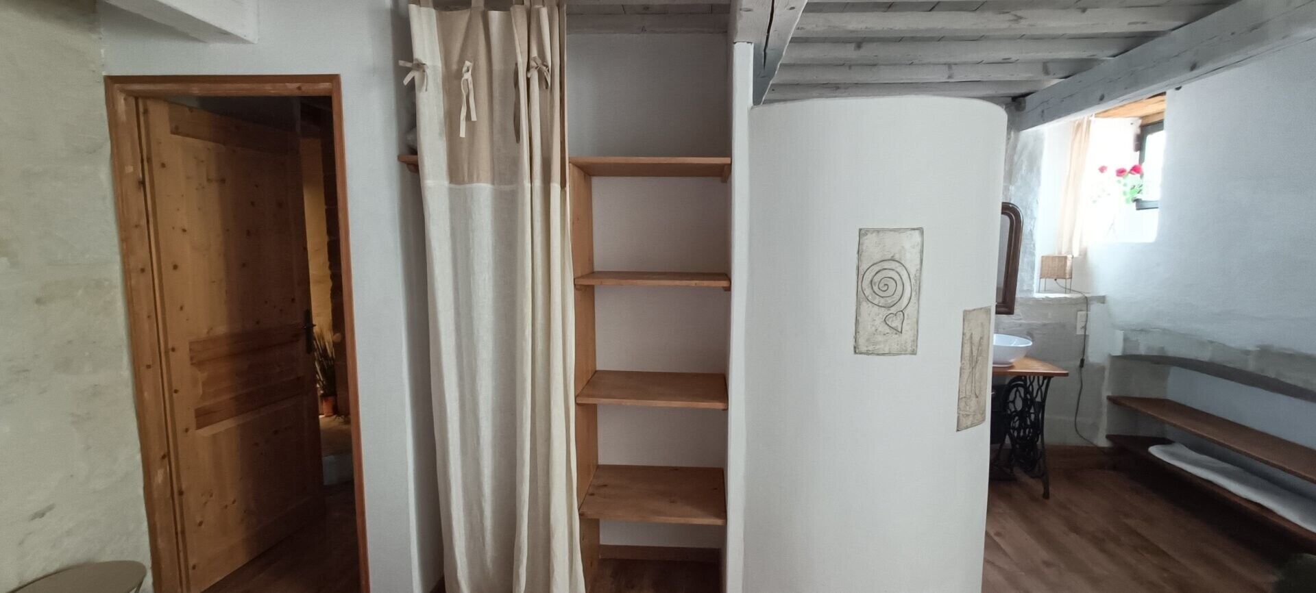 Une pièce avec une porte en bois, un rideau blanc, une étagère en bois, une machine à coudre et une fenêtre donnant sur des fleurs.