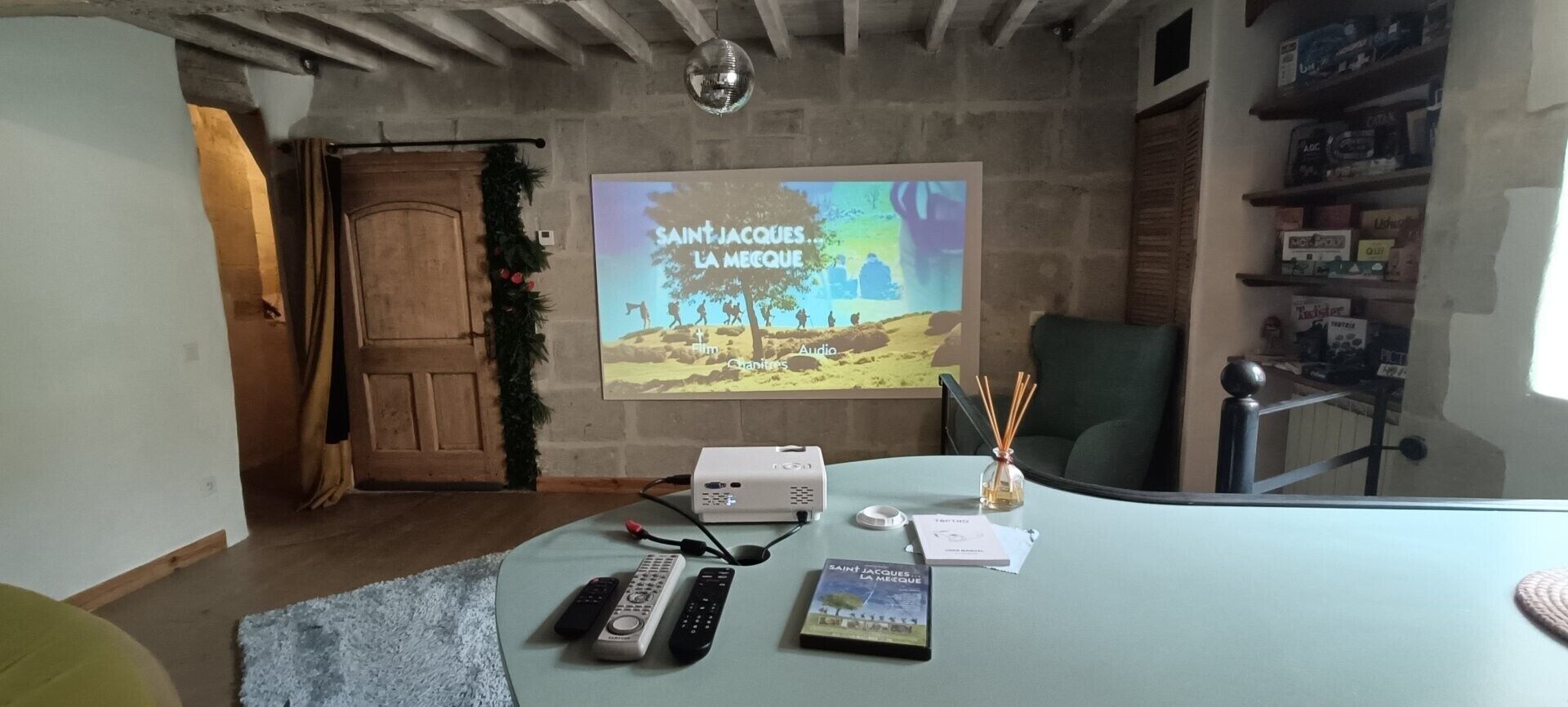 Een kamer met een projectiescherm waarop 'SAINT-JACQUES LA MECOQUE' staat, een houten deur, een groene stoel, een tafel met afstandsbedieningen, een boek en een decoratieve guirlande.