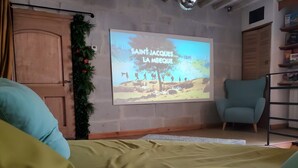 Une pièce dotée d’un écran de projection sur lequel est affiché le titre « SAINT-JACQUES LA MECOUE », d’une porte en bois, d’un fauteuil bleu et d’une étagère contenant divers objets.