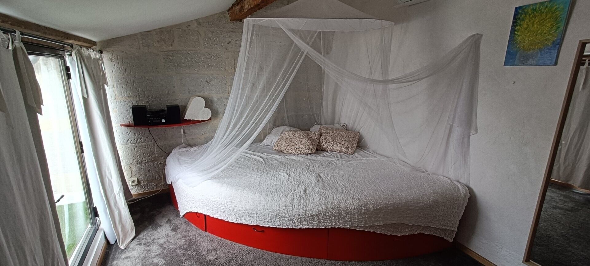 Ein Zimmer mit einem Himmelsbett, einem kleinen Regal mit einem Radio und einer herzförmigen Dekoration sowie einem Bild an der Wand.