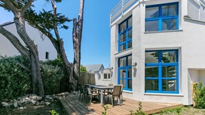 A modern building with blue-framed windows, a wooden deck, and a dining area with chairs.