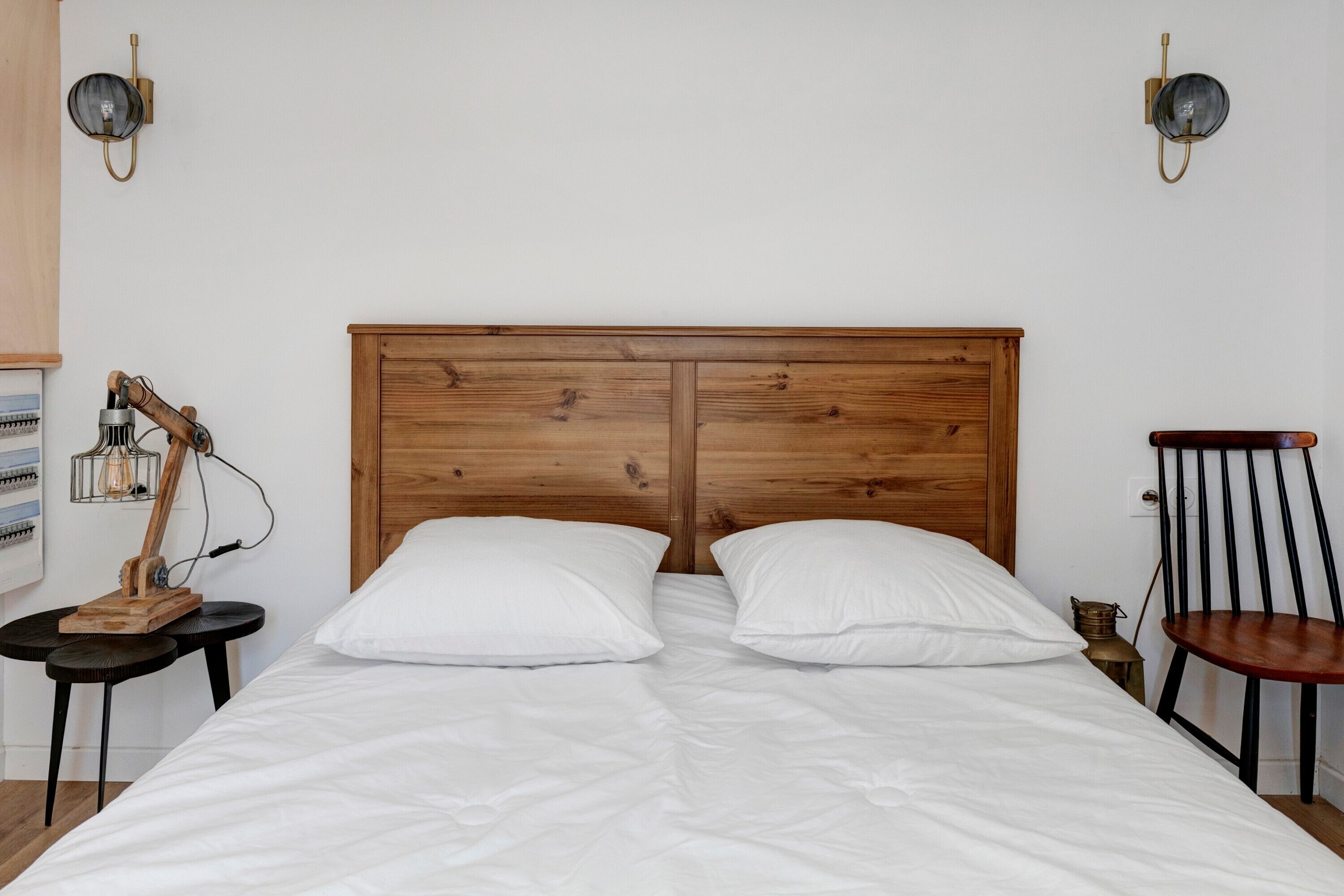 Een net opgemaakt bed met witte lakens, een houten hoofdbord, twee wandlampen en een klein tafeltje met een lamp.