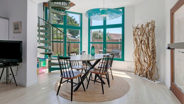 A modern living room with a dining table, chairs, a TV, a staircase, and large windows.