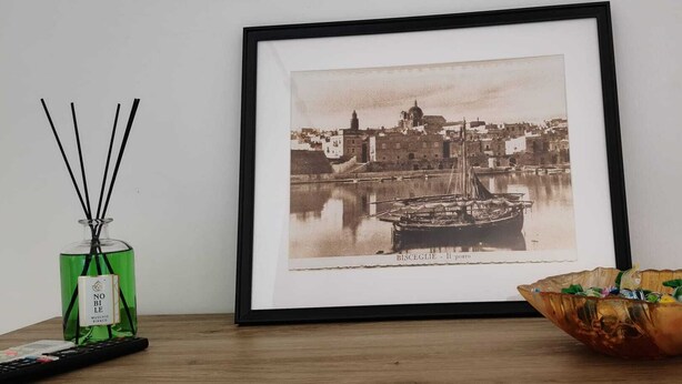 Una fotografía enmarco de un paisaje urbano histórico que muestra un barco, una botella verde con palitos negros y un cuenco decorativo sobre una mesa de madera.