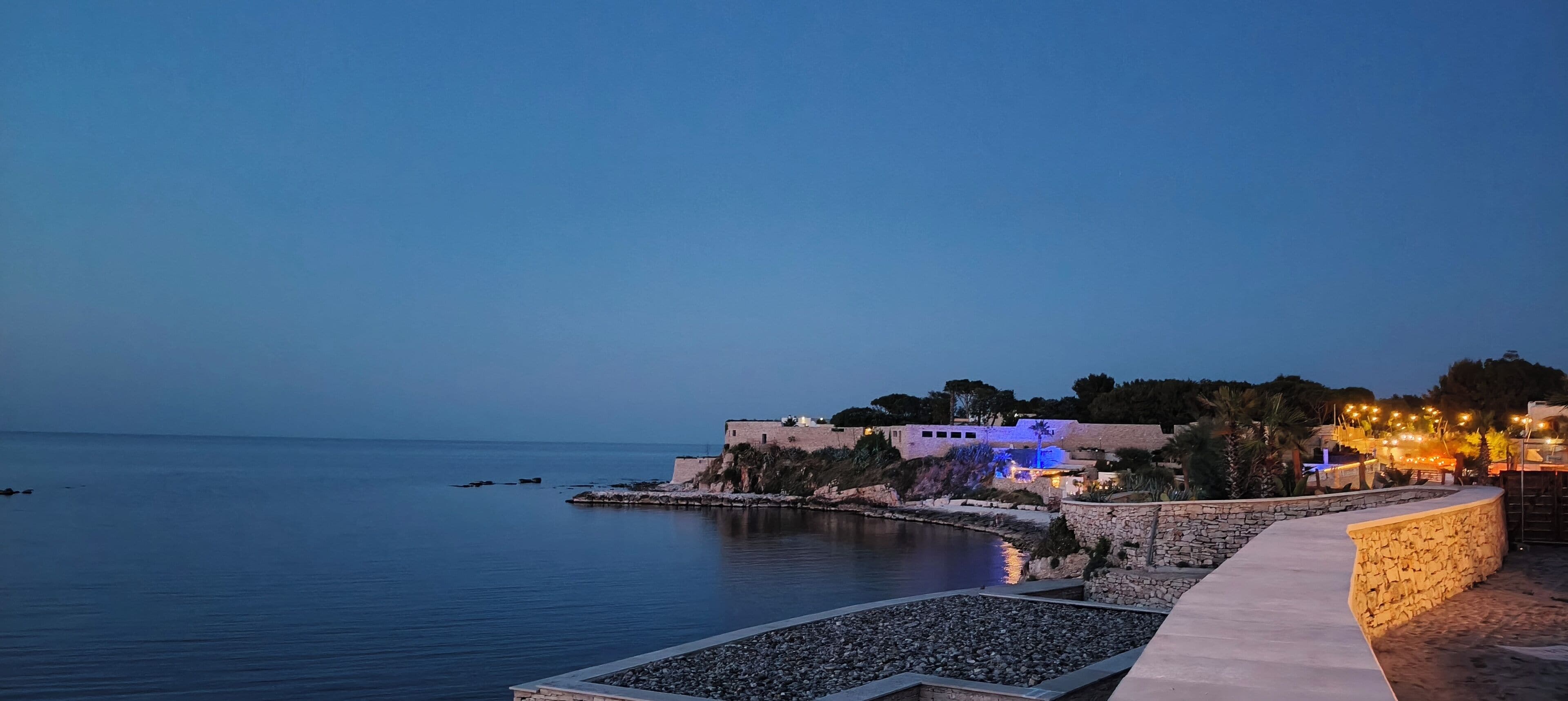 A coastal property with a stone pathway leading to a water body, a building with illuminated windows, and a clear view of the sea.