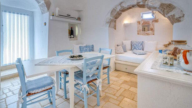 Un espace repas comprenant une table, des chaises et un banc. On y trouve une arche en pierre, une fenêtre et une étagère avec divers objets.