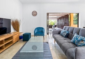 A living room with a TV, blue armchair, grey sofa, and a glass coffee table. - Spacious 3 Bed Room House, Pool Ideal Families & Groups, Secure & Central (Durban North)