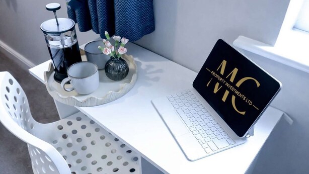 A white desk with a laptop displaying "MC Property Investments Ltd" and a tray with a coffee maker, mug, and flowers.