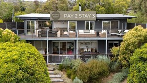 Ein modernes, zweistöckiges Gebäude mit einem Balkon, einem Schild mit dem Hinweis auf ein 'PAY 2 STAY 3' Angebot und üppiger Bepflanzung darum herum.