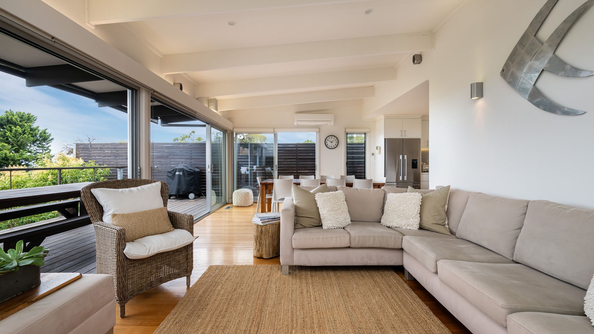 Un vaste espace de vie comprenant un grand canapé, un fauteuil en osier et une table à manger. Une porte-fenêtre coulissante donne accès à un balcon équipé d’un barbecue.