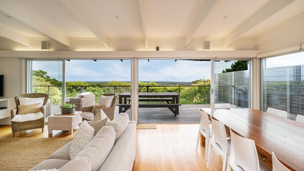 Ein großzügiger Wohnbereich mit einem großen Fenster und Blick auf die Landschaft. Es gibt ein Sofa, einen Esstisch und einen Korbstuhl.