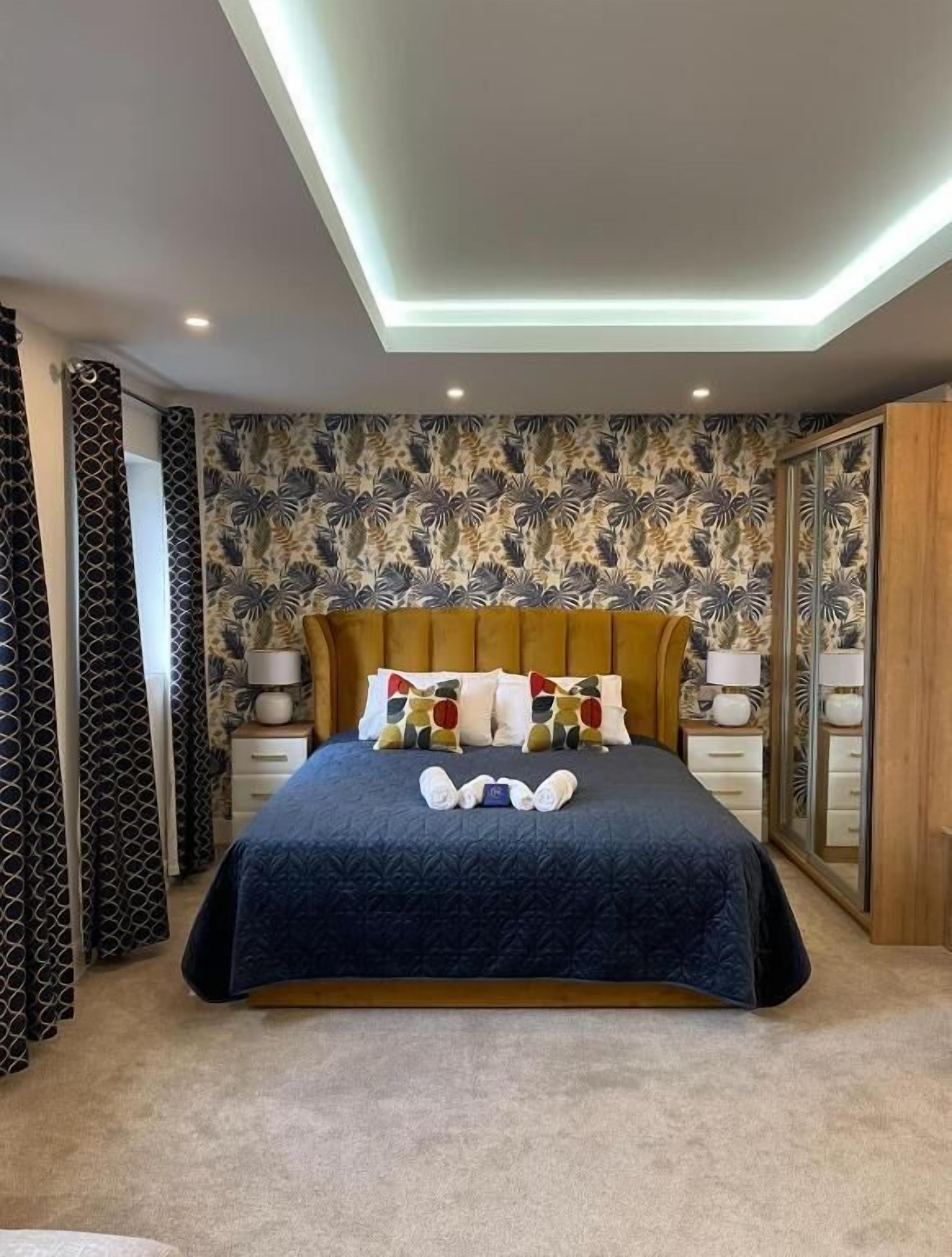 A neatly made bed with a dark blue bedspread, a wooden headboard, and colorful pillows. There are two bedside tables with lamps and a mirror. The room has a patterned wallpaper and a ceiling light.
