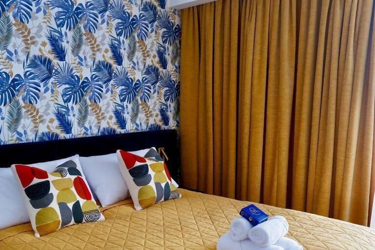 Una habitación de hotel con cama, dos almohadas, colcha acolchada, cabecera y cortina.
