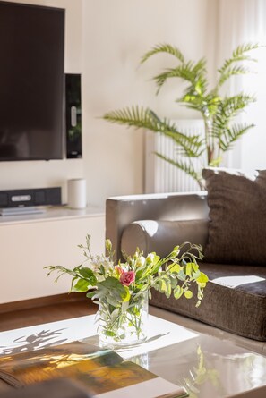 Een woonkamer met een bank, een bijzettafeltje met een bloevas en een televisie aan de muur.