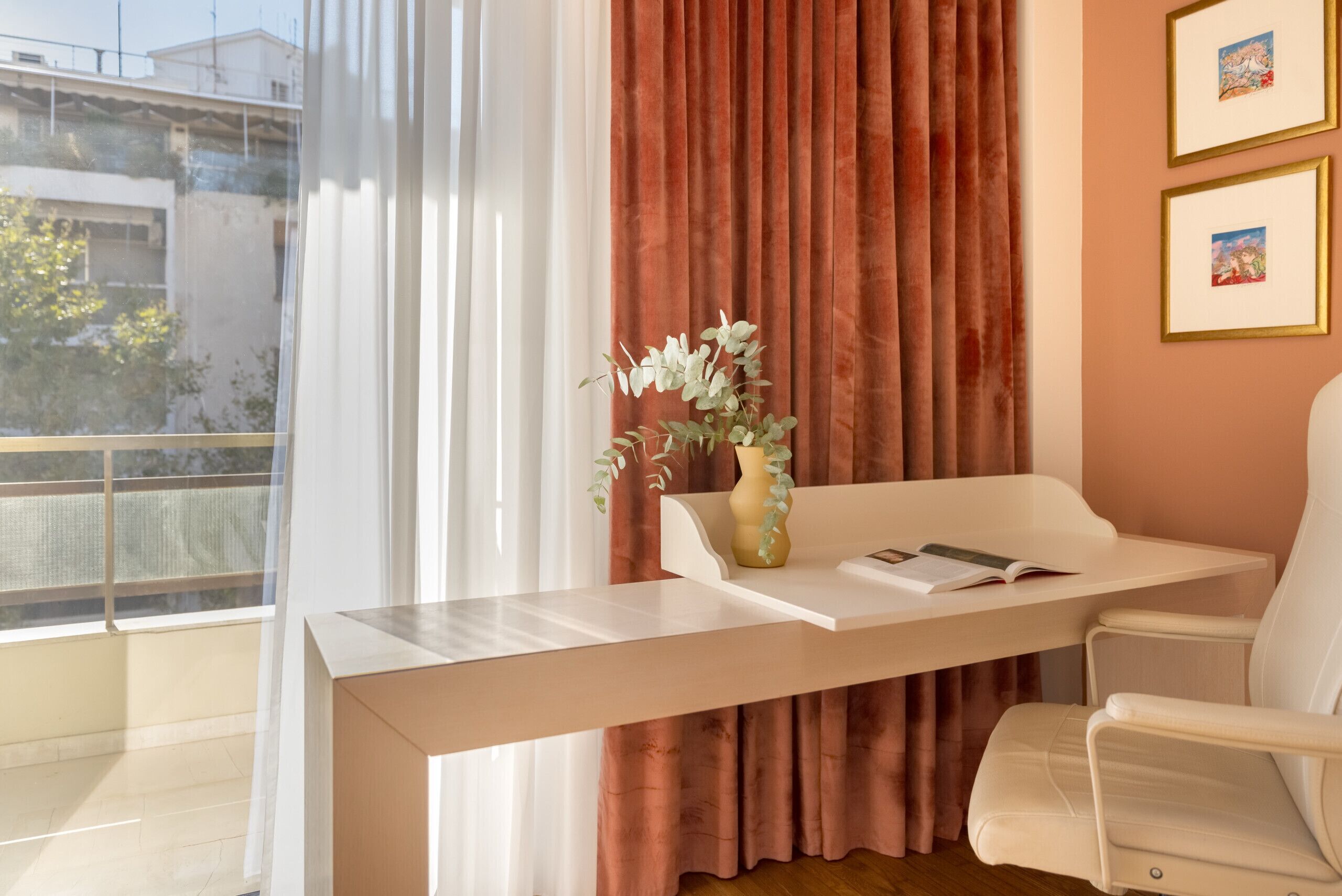Une pièce moderne avec un bureau blanc, un vase orné de feuillage vert, une chaise blanche et une fenêtre avec des rideaux légers.