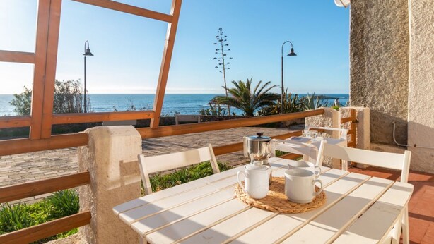A table with a white tablecloth, a white mug, and a white teapot on a woven placemat, overlooking a beach with a clear sky.
