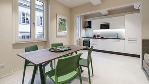 A modern kitchen with white cabinets, a dining table with green chairs, and a window with blinds. - Palazzo Accademia-brunelleschi-1 (Florence)