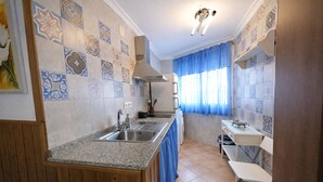 Una cocina con encimera de granito, pileta de acero inoxidable y estufa. Hay una ventana con cortinas azules y una pequeña mesa con una pileta.