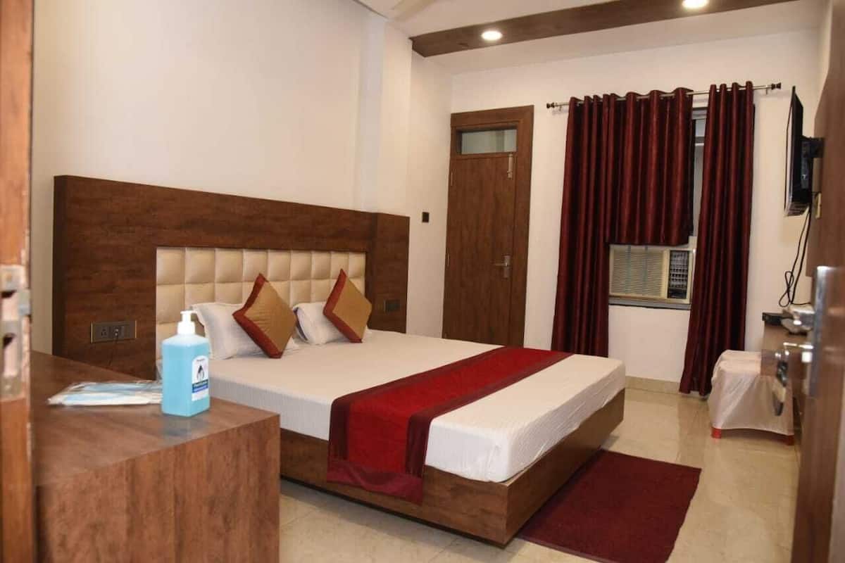 Ein Hotelzimmer mit Holz-Kopfteil, einem Bett mit weißen Bettwäsche und roten Akzenten, einem Schreibtisch mit Handdesinfektionsmittel und einem Fenster mit Vorhängen.