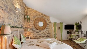 A bedroom with a stone wall, a round mirror, a bed with leaf-patterned pillows, a bedside lamp, a small table with a plant, and a TV mounted on the wall. - Bed and Breakfast 'Chambre Nature' with Shared Garden and Wi-Fi (Saint-Malô-du-Bois)